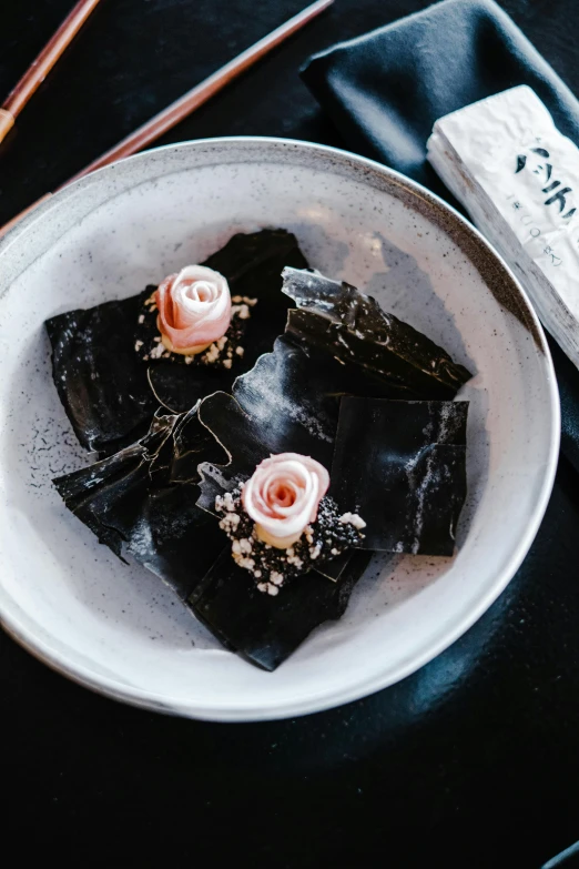 a plate of food on a table with chopsticks, black roses, floating kelp, wrapped in black, pink petals