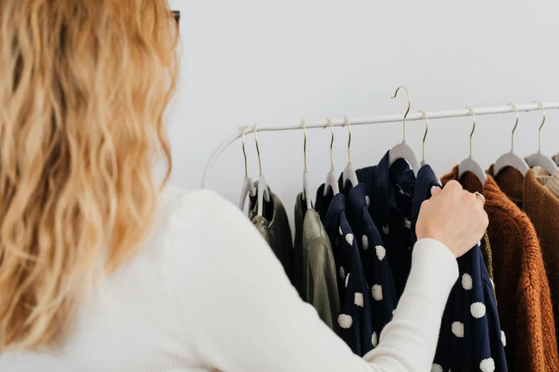 a woman standing in front of a rack of clothes, by Nicolette Macnamara, trending on pexels, private press, with a white background, inspect in inventory image, back towards camera, linsey levendall