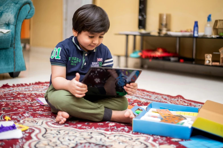 a young boy sitting on the floor playing with a tablet, by Julia Pishtar, hurufiyya, faridah malik, playing board games, with book of science, multi-dimensional