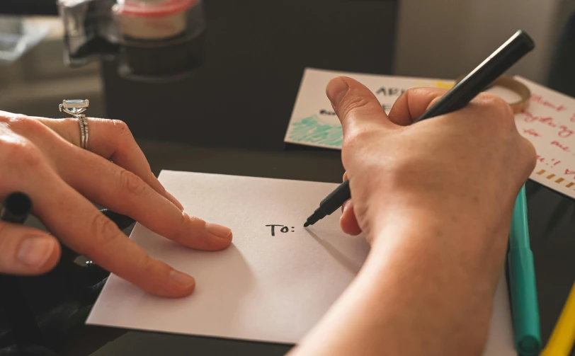 a person writing on a piece of paper with a pen, by Sam Black, pexels contest winner, letterism, cards, hand on table, close - up photo, thumbnail