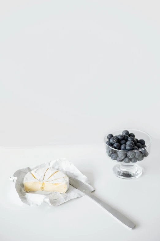 a piece of cheese sitting on top of a table next to a bowl of blueberries, visual art, white space, grey, glassware, uncropped