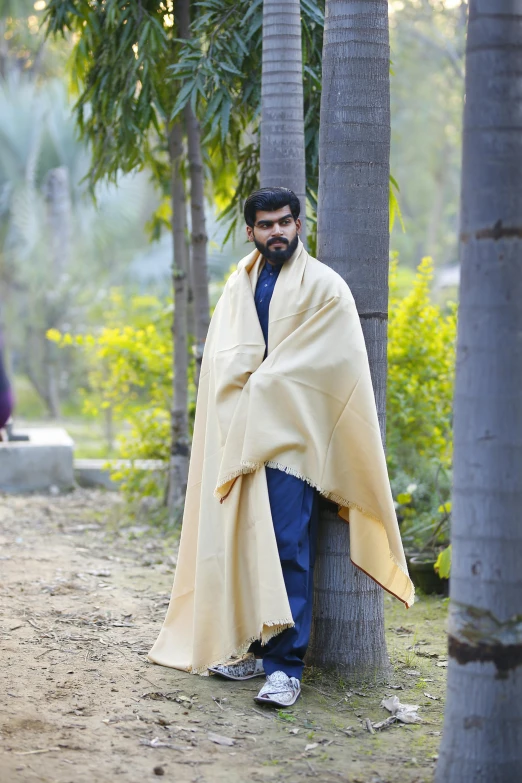 a man wrapped in a blanket standing next to a tree, a portrait, inspired by Kailash Chandra Meher, pexels, at a fashion shoot, wearing wheat yellow gauze, post graduate, taken in zoo