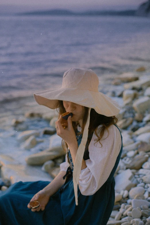 a woman sitting on a rocky beach talking on a cell phone, inspired by Oleg Oprisco, unsplash, white straw flat brimmed hat, smokes, bluish and cream tones, ulzzang