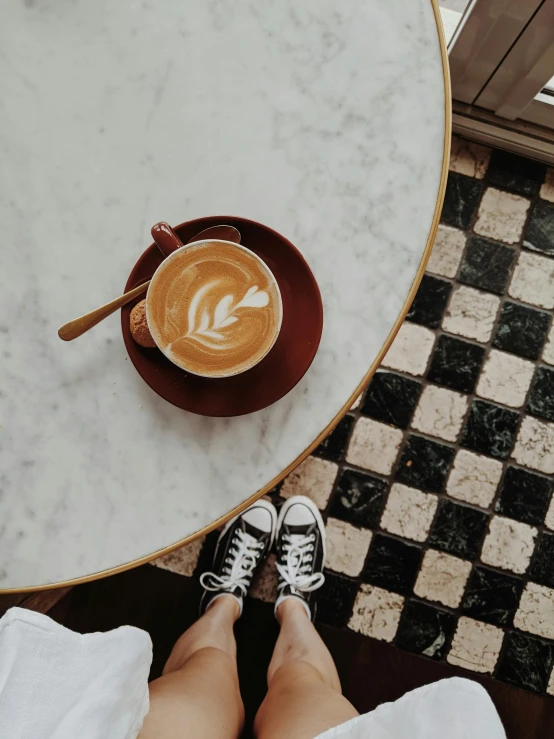 a person sitting at a table with a cup of coffee, wearing white sneakers, on a mosaic marble floor dais, instagram story, aussie baristas