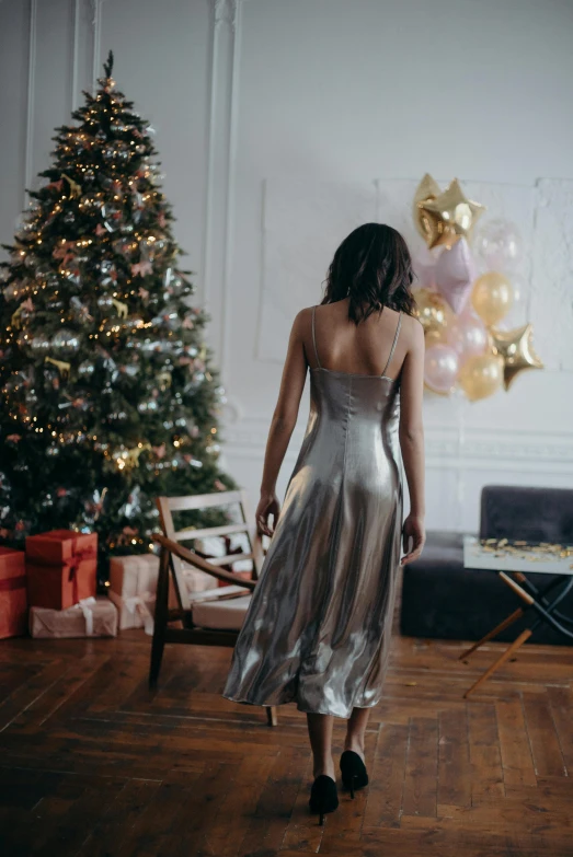 a woman standing in front of a christmas tree, happening, wearing silver dress, back towards camera, at home, promo image