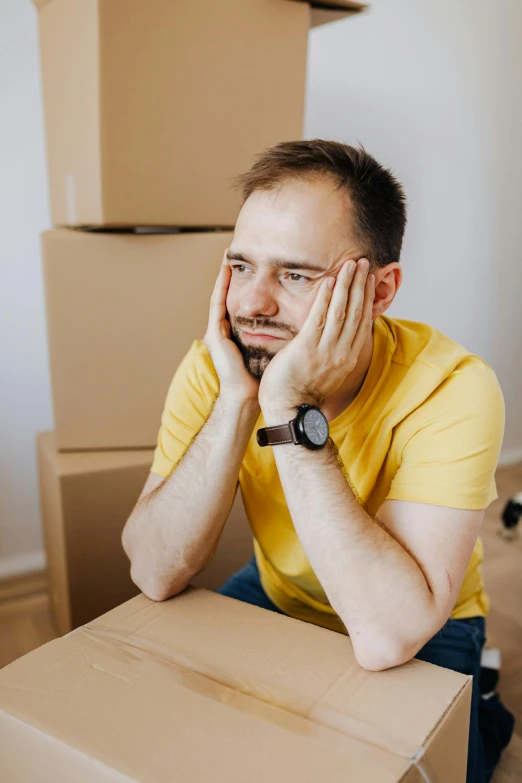 a man sitting on top of a cardboard box, looking exhausted, wearing a modern yellow tshirt, lean man with light tan skin, headshot profile picture