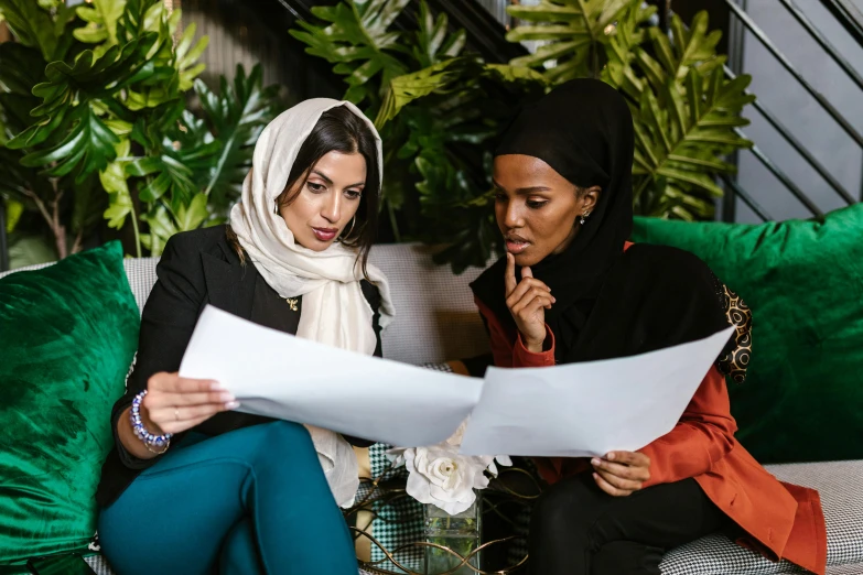 two women sitting on a couch looking at a piece of paper, hurufiyya, white hijab, thumbnail, professional image, contain