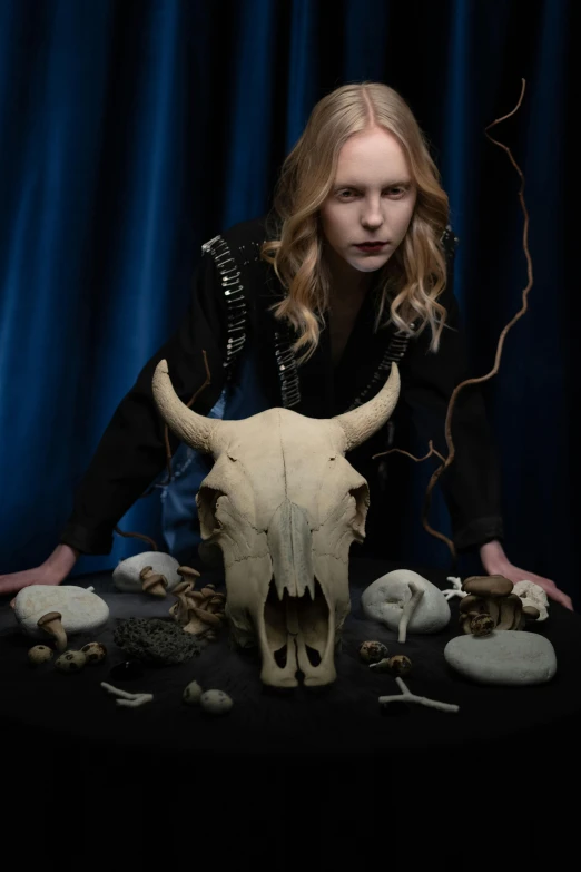 a woman sitting on top of a table next to a cow skull, wearing gothic accessories, resembling a mix of grimes, johannen voss, shamanistic dark blue clothes