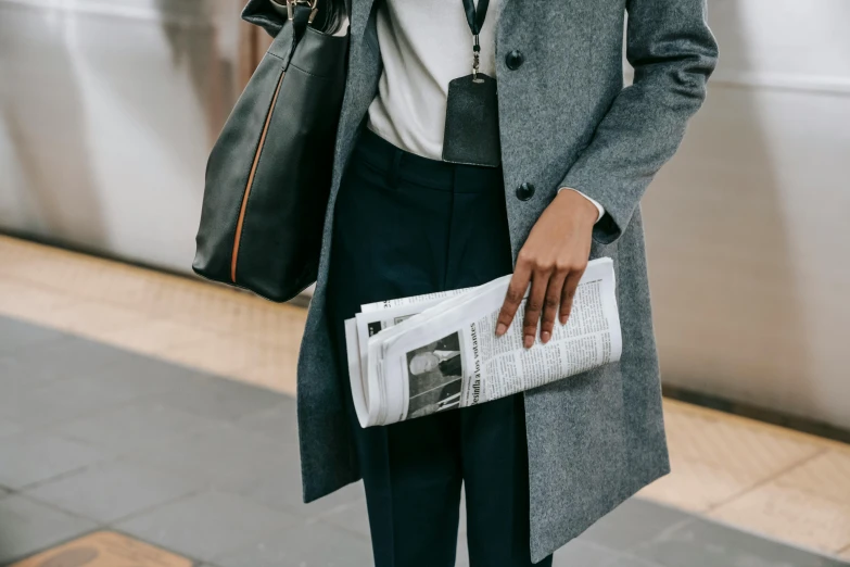 a woman standing next to a train holding a newspaper, by Nina Hamnett, trending on unsplash, trench coat and suit, holding a briefcase, grey clothes, news feed