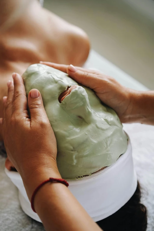 a woman getting a clay mask on her face, hands shielding face, large head, a green, bum