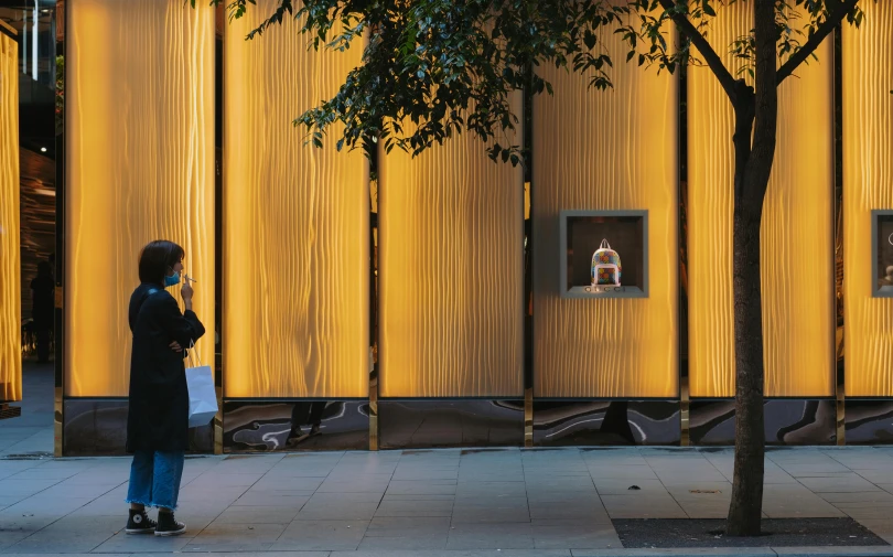 a woman standing on a sidewalk next to a tree, a hologram, inspired by Christo, unsplash contest winner, interactive art, yellow awning, in front of a carved screen, gold and luxury materials, hugh kretschmer