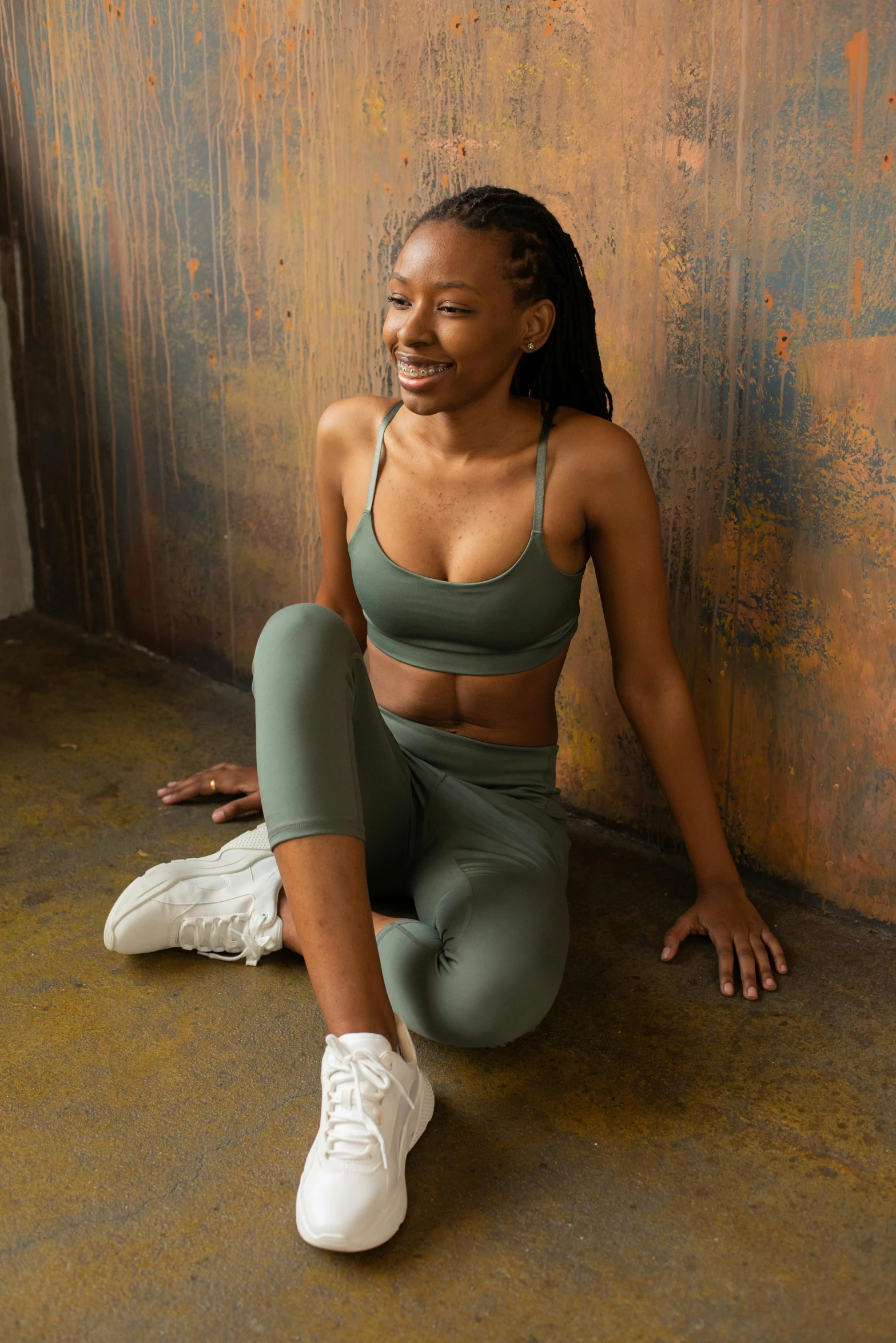 a woman in a sports bra top and leggings sitting on the floor, by Ella Guru, muted green, ( ( dark skin ) ), smiling down from above, leaning on the wall