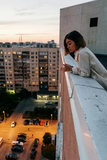 a woman standing on top of a building next to a parking lot, inspired by Elsa Bleda, pexels contest winner, happening, soviet apartment building, checking her phone, gazing off into the horizon, high angle