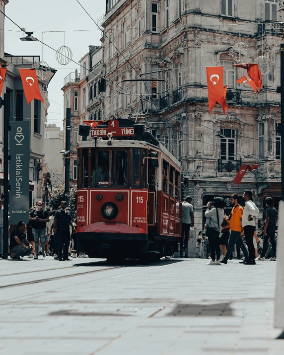 a red trolley traveling down a street next to tall buildings, pexels contest winner, ottoman empire era, gay pride, background image, a group of people