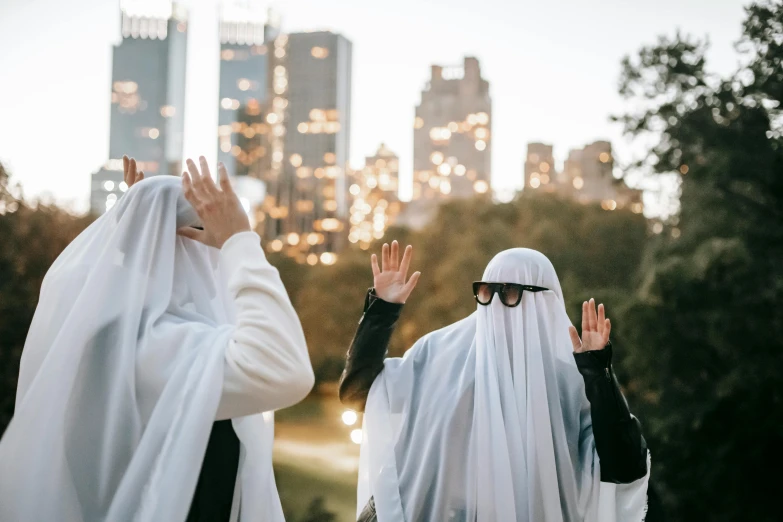 two people dressed in white standing next to each other, by Julia Pishtar, unsplash contest winner, hurufiyya, halloween theme, city skyline in the background, waving robe movement, posing in disguise among humans