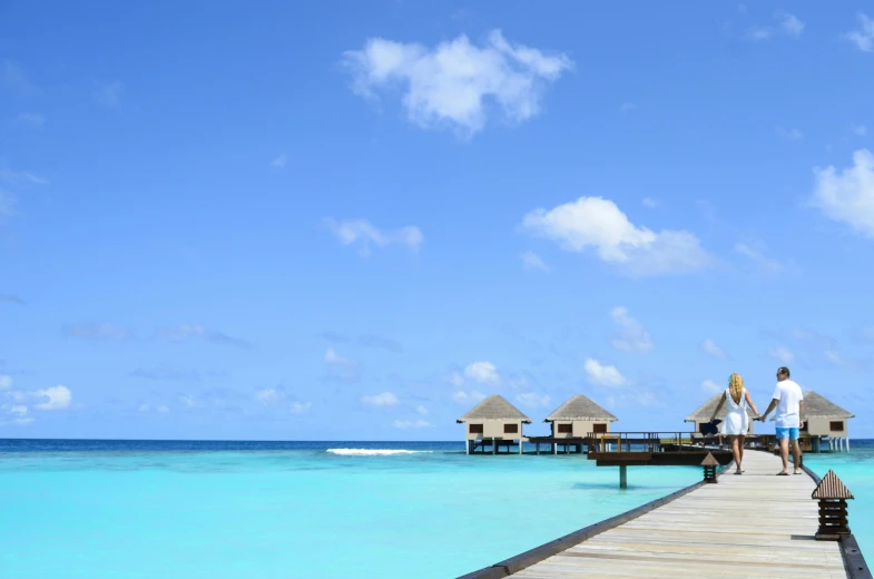 a man and a woman walking on a pier over the ocean, an album cover, trending on unsplash, maldives in background, cloudless blue sky, huts, thumbnail