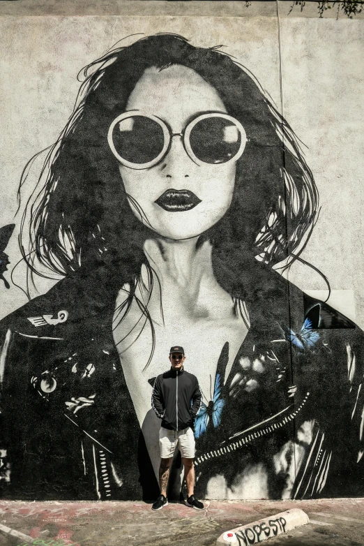 a man standing in front of a mural of a woman, poster art, by Micha Klein, trending on pexels, street art, dressed in biker leather, sunglasses, adi granov, dope