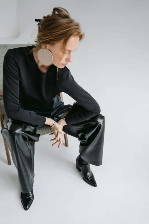 a woman sitting on top of a wooden chair, by Nina Hamnett, trending on unsplash, bauhaus, black leather shiny jeans, black turtle neck shirt, huge earrings, androgyny