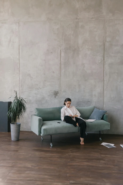 a woman sitting on a couch reading a book, inspired by Constantin Hansen, minimalism, rough concrete walls, office furniture, muted green, connectivity