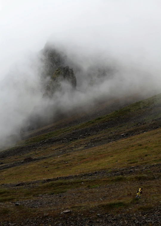 a man flying a kite on top of a lush green hillside, by Hallsteinn Sigurðsson, hurufiyya, uneven dense fog, with jagged rocks & eerie, seen from a distance, ceremonial clouds