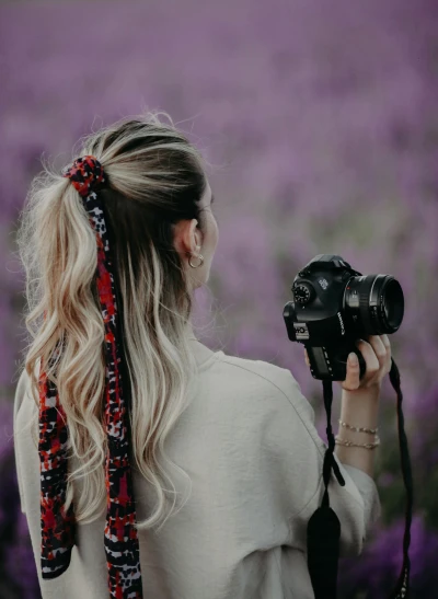 a woman holding a camera in front of a field of purple flowers, unsplash contest winner, ribbon in her hair, long ponytail, red rose in hair, photography]