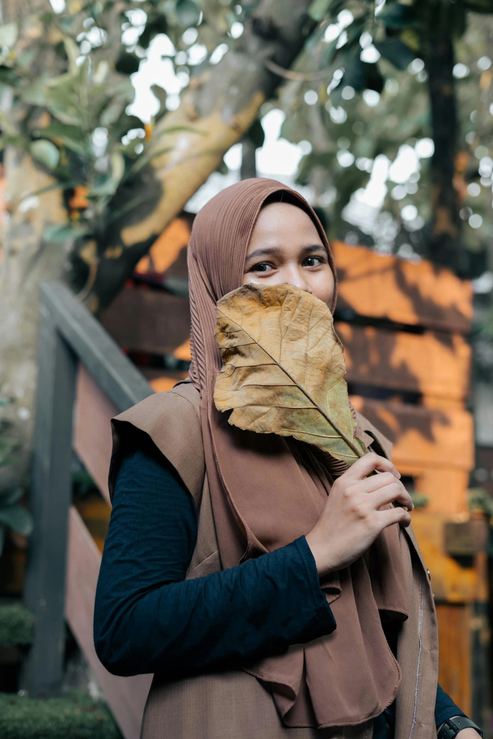 a woman holding a leaf in front of her face, by Basuki Abdullah, pexels contest winner, hurufiyya, wearing a brown, wearing a scarf, al fresco, a wooden