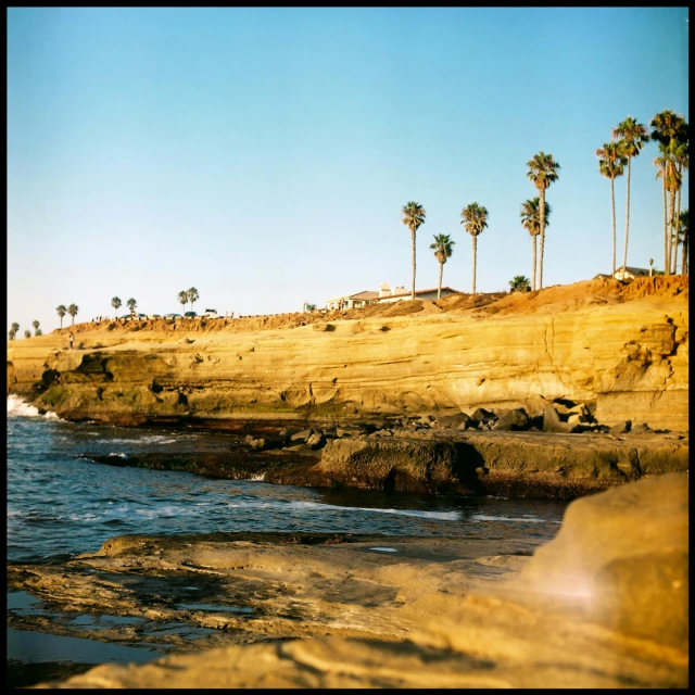 a man riding a surfboard on top of a sandy beach, a polaroid photo, by Andrew Domachowski, unsplash, photorealism, trees and cliffs, a palm tree, shiny layered geological strata, the city of santa barbara