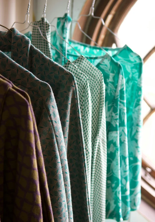 clothes hanging on a rack in front of a window, a silk screen, by Elizabeth Charleston, unsplash, renaissance, loose - fitting blouses, jade green, subtle patterns, wearing a colorful men's suit