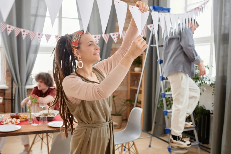 a woman hanging bunting flags on a string in a living room, pexels contest winner, arbeitsrat für kunst, long dreadlocks, people at work, in a white boho style studio, at a birthday party