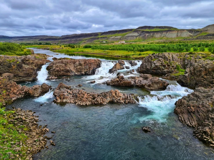 a river running through a lush green valley, by Þórarinn B. Þorláksson, pexels contest winner, hurufiyya, waterfalls in the background, with lots of dark grey rocks, thumbnail, photo for magazine