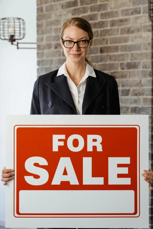 a woman holding a for sale sign in front of a brick wall, shutterstock, photorealism, wearing a suit and glasses, in house, inside a grand, low density