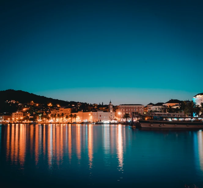 a large body of water next to a city at night, pexels contest winner, romanticism, picton blue, sunny sky, reflections in copper, croatian coastline