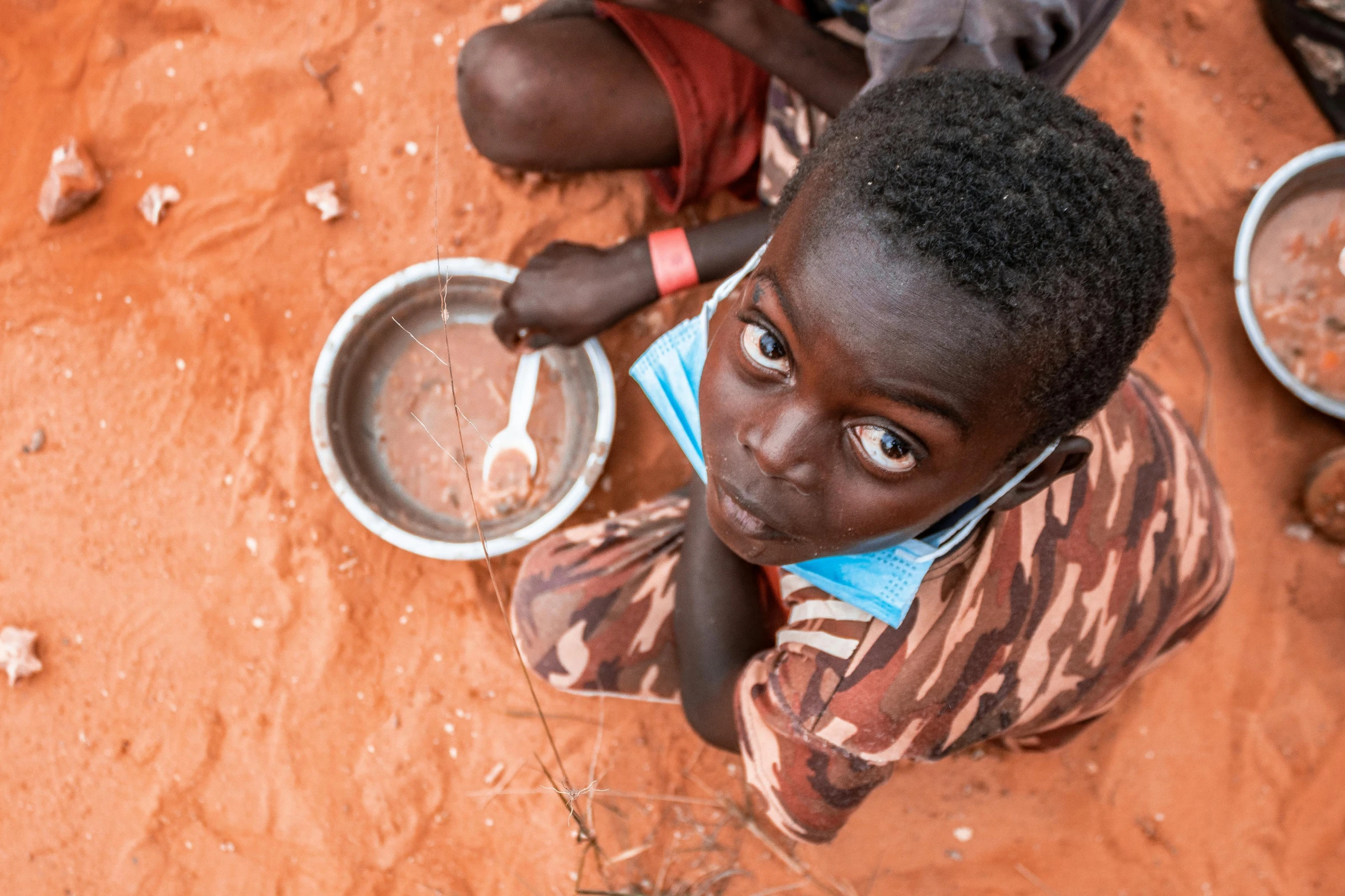 a young boy sitting on the ground eating food, by Daniel Lieske, hurufiyya, red sand, looking down on the camera, soup, emmanuel shiru