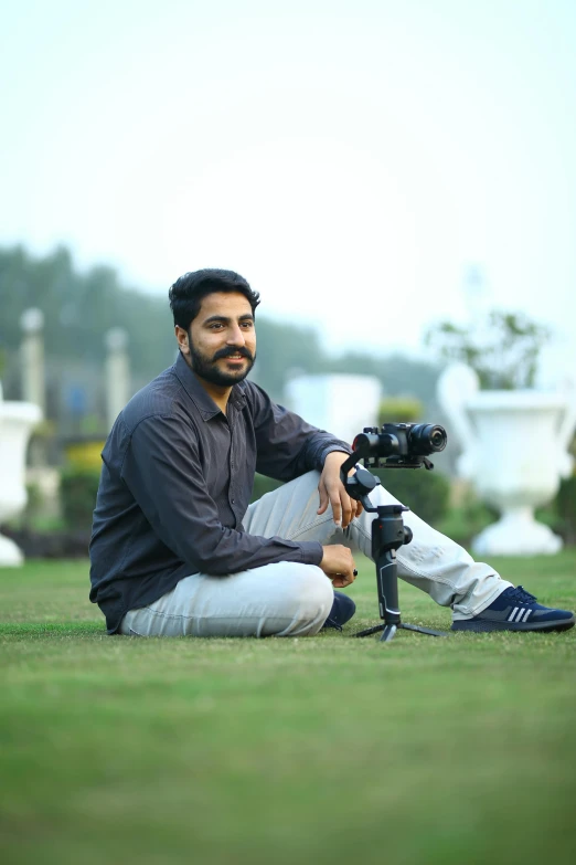 a man sitting on the ground with a camera, by Riza Abbasi, in a scenic background, a portrait of rahul kohli, professional profile picture, slightly pixelated