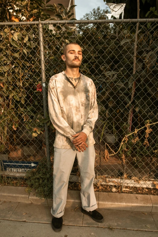 a man standing in front of a chain link fence, an album cover, inspired by Elsa Bleda, renaissance, blood stains on shirt, wearing cargo pants, brown buzzcut, tie-dye