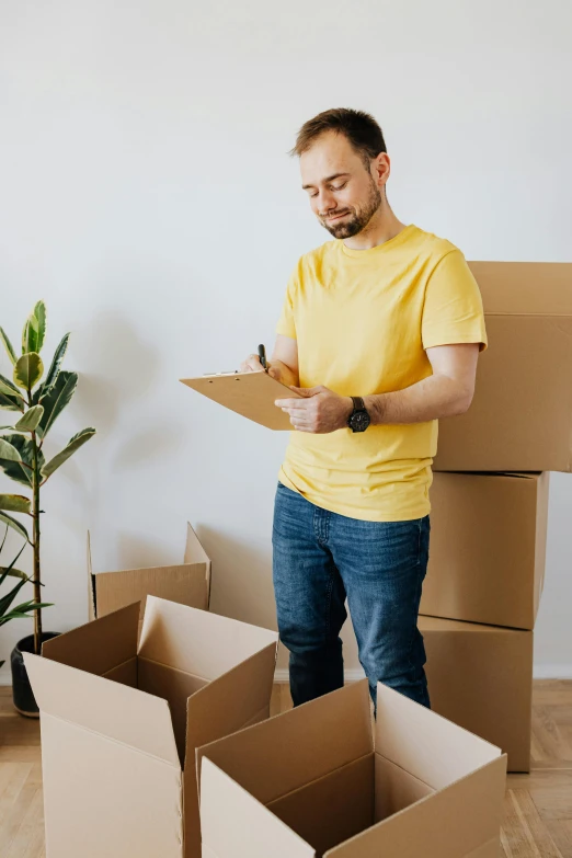 a man standing next to a pile of cardboard boxes, writing on a clipboard, yellow theme, australian, on high-quality paper