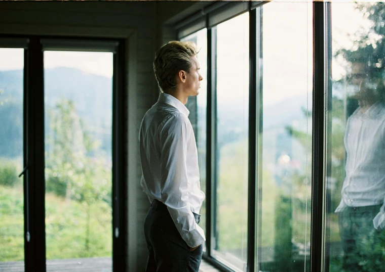 a man standing in front of a window looking out, by Julia Pishtar, in karuizawa, profile image, groom, concerned