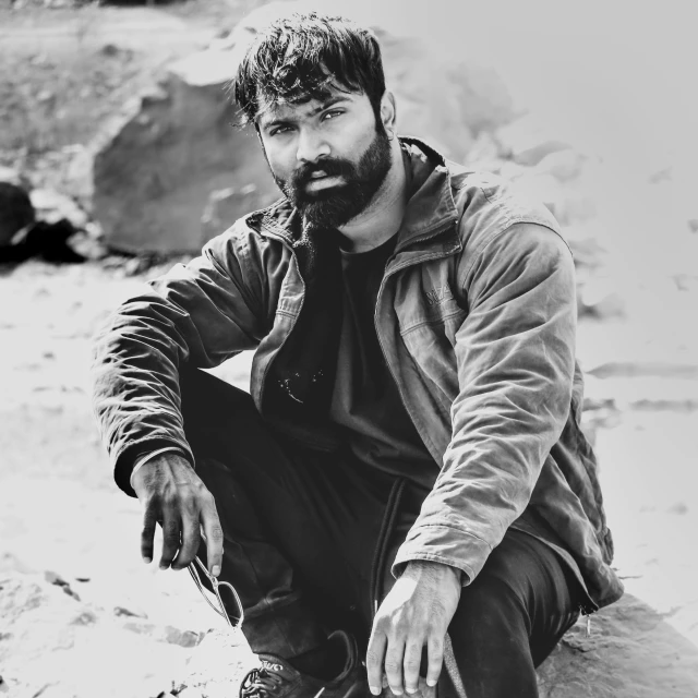 a black and white photo of a man sitting on a rock, a sketch, hurufiyya, very attractive man with beard, wearing ripped dirty flight suit, a portrait of rahul kohli, autumn season