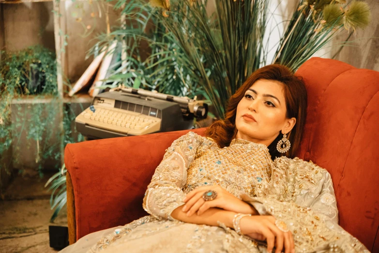 a woman sitting in a chair next to a typewriter, an album cover, inspired by Lubna Agha, pexels, qajar art, gold suit, lounging on expensive sofa, wearing elegant jewellery, cinematic outfit photo