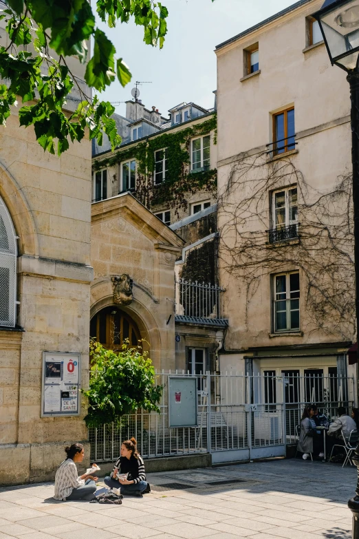 a group of people sitting on the ground in front of a building, paris school, archways made of lush greenery, shady alleys, library, whitewashed buildings