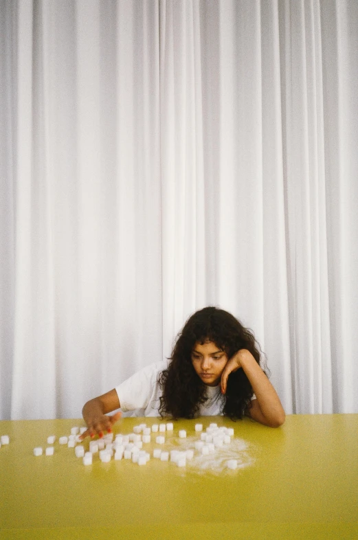 a woman sitting at a table with marshmallows in front of her, inspired by Elsa Bleda, imaan hammam, board games on a table, in front of white back drop, curls on top