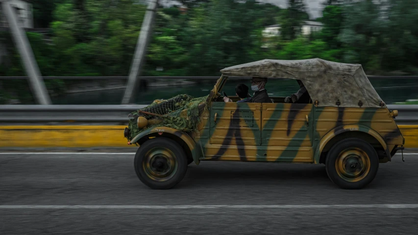 a military vehicle driving down a street next to a forest, a colorized photo, unsplash, photorealism, manila, camouflaged gear, driving a hotrod, tigerman