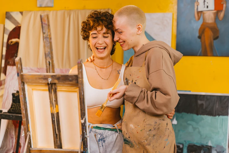 a couple of people standing in front of a painting, by Julia Pishtar, pexels contest winner, arbeitsrat für kunst, lesbian embrace, both laughing, stands at her easel, non binary model