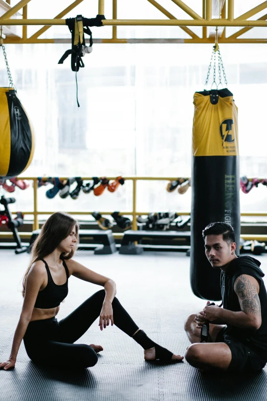 a man and a woman sitting on the ground in a gym, by Robbie Trevino, punching in a bag, black and yellow color scheme, manila, confident