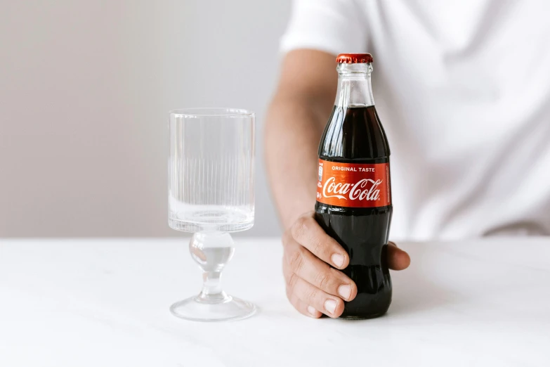 a person holding a bottle of coke next to a glass, on a marble pedestal, product shot, traditional medium, glassware