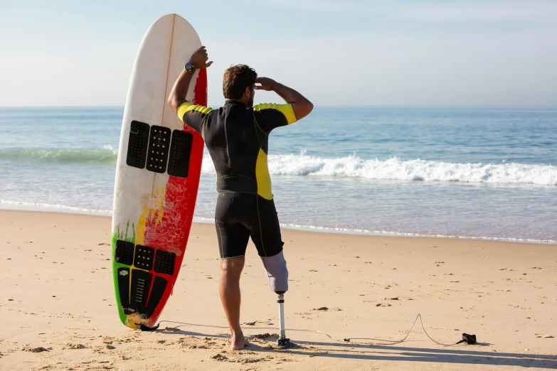 a man standing on a beach holding a surfboard, prostheses, injured, multicoloured, rocha