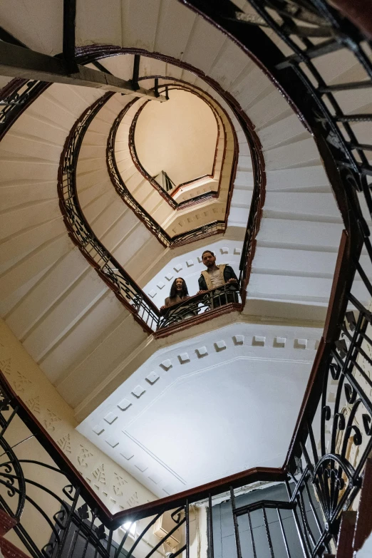 a couple of people standing at the top of a spiral staircase, vienna secesion style, branching hallways, head straight down, back towards camera