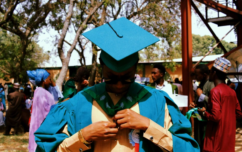 a man in a cap and gown adjusts his tie, by Ingrida Kadaka, unsplash, sea - green and white clothes, samburu, at college, with teal clothes