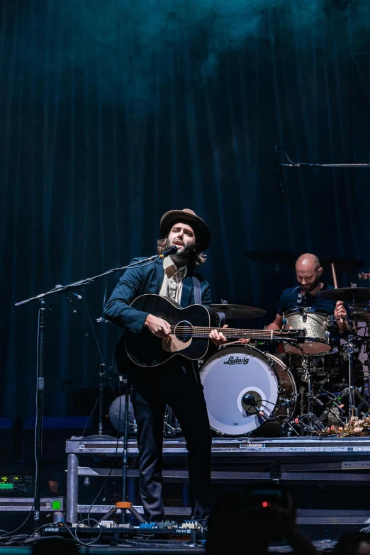 a man standing on top of a stage holding a guitar, tipping his fedora, matt berry, zoomed out, wilderness ground