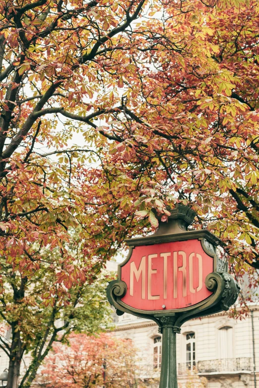 a red metro sign sitting on the side of a road, inspired by Oleg Oprisco, trending on unsplash, art nouveau, maple trees with fall foliage, paris hotel style, square, station